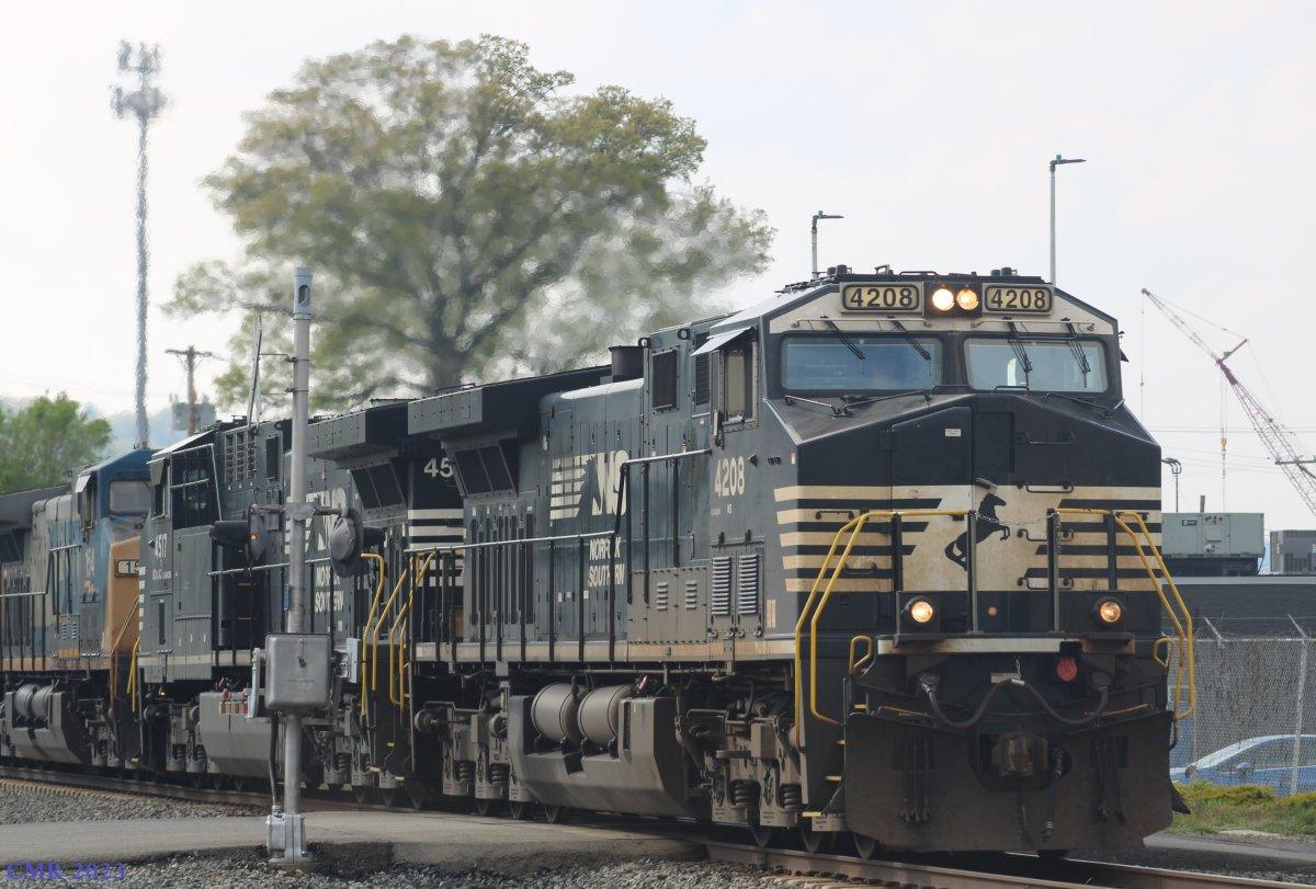 A Norfolk Southern freight train led by locomotive 4208 in black‑and‑white livery, followed by units 4517 and another locomotive further back. The train moves along a track bordered by industrial structures including a crane, utility poles, and a fenced facility, with trees and buildings in the background.